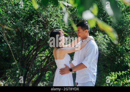 Passionate asian couple in white clothes standing in a bush in a park, away from the eyes of others. Looking at each other, holding one another. Stock Photo