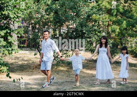Joyful asian family in white clothes having a walk on a grass in a park, enjoying last warm days of the early fall. Slowly strolling between trees, ho Stock Photo
