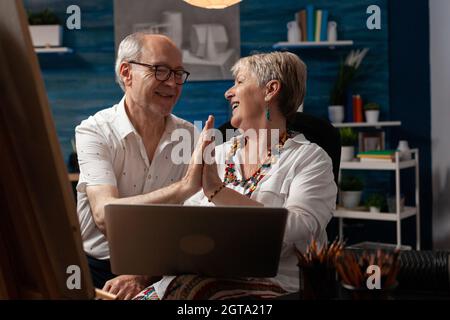 Aged couple of artists highfive while using laptop for design inspiration in workshop space. Caucasian elder man and woman feeling cheerful and working on drawing for fine art project Stock Photo