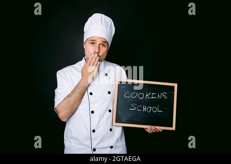 Cook dressed in a jacket and white hat puts his hand to his mouth and holds a blackboard that reads: Cooking School on a black background. Restaurant, Stock Photo