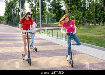 Three Young Girl Friends On Vacation Having Fun Driving Electric Scooter Through The City Park.