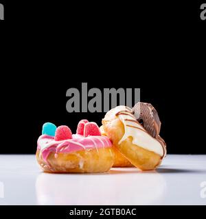 Photograph of a composition of 3 donuts decorated with colored chocolate, cookies and jelly beans.The photo is taken on a white table and a black back Stock Photo