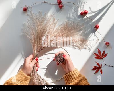 Hands making dried floral wreath from dry pampas grass and Autumn leaves. Hands in sweater with manicured nails tie decorations to metal frame. Flat Stock Photo