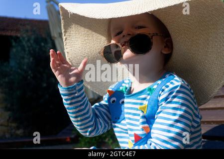 Cute little toddler wearing an oversized sun hat and sunglasses relaxing in the sun Stock Photo