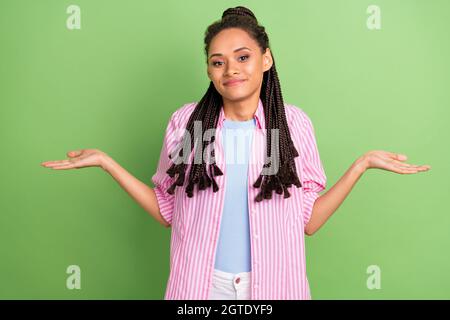 Photo of clueless funny dark skin woman dressed striped shirt shrugging shoulders smiling isolated green color background Stock Photo