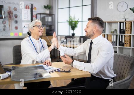 Happy man in business wear giving high five to female doctor at medical cabinet. Successful recovery after professional treatment. Medicine concept. Stock Photo