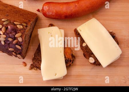 on a wooden board made of light wood lie two sandwiches with cheese next to it lies a sausage and a loaf of bread from which they cut it off. Stock Photo