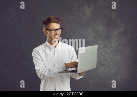 Smiling man stands on gray background and types text on laptop which he holds in his hands. Stock Photo