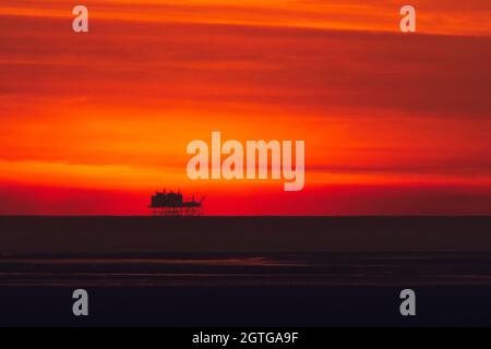 Silhouette of an oil rig or platform on a distant horizon against dramatic orange sky at sunset. Theme or concept of fossil fuels, sunset industry Stock Photo