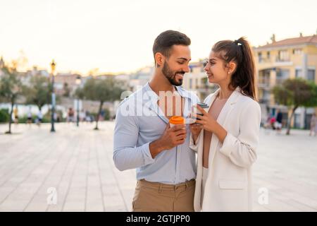 Cheerful ethnic stylish couple with takeaway coffee standing close to each other on city square in evening and enjoying stroll Stock Photo