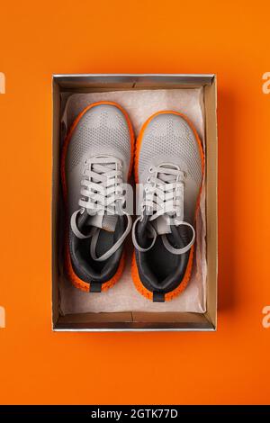 Pair of new gray mesh fabric sneakers in the open box on the bright orange background. Modern textile trainers with grooved orange sole for sport. Stock Photo