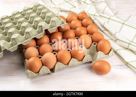 One brown chicken egg out of the full cardboard tray on a white wooden table. Fresh organic hen eggs with brown shell in the biodegradable carton cont Stock Photo