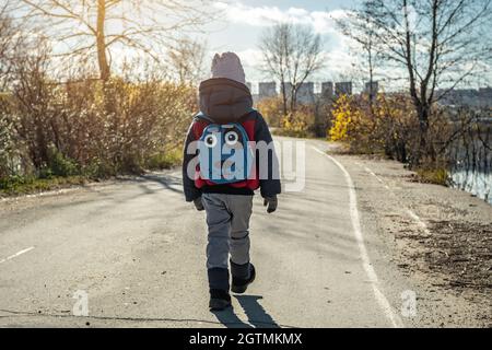 A child boy hiking with a small backpack is walking along the road outside the city on the background of the autumn landscape Stock Photo