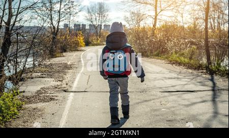 A child boy hiking with a small backpack is walking along the road outside the city on the background of the autumn landscape Stock Photo