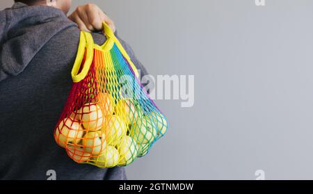 A man holds in his hands a reusable cotton shopping string bag, a multicolored rainbow with green apples inside. On a gray background. Zero waste conc Stock Photo