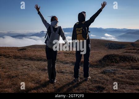 Hiking adventure healthy outdoors people standing talking. Couple enjoying view above clouds on trek. young woman and man in nature wearing hiking backpacks and sticks. Stock Photo