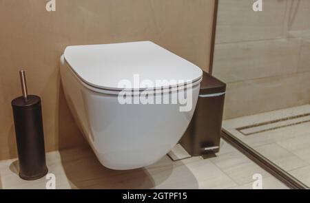 Toilet interior, ceramic white toilet bowl and black brush. Modern bathroom accessories on brown walls and tiled floor background. Stock Photo