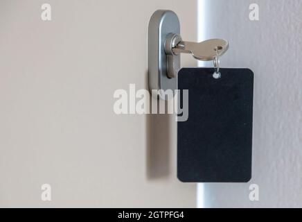 Door key with blank label in the keyhole. Steel key ring, room key and empty black tag for text or number close up view. House, hotel or motel room op Stock Photo