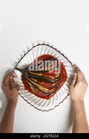 Human hands putting delicious dish made of eggplant with tomatoes and sauce on a plate Stock Photo