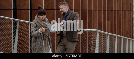 Handsome businessman with female colleague using laptop and smiling on a terrace outside office, working. Grey haired man in autumn brown trendy style Stock Photo
