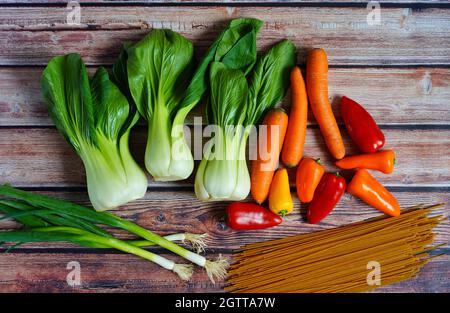 Fresh spring onions, pak choi, carrots, bell pepper and wholemeal noodles on a rustic wooden table. Preparation for healthy home cooking. Stock Photo
