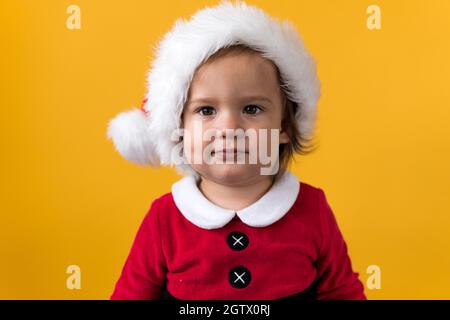 Smiling Portraite Cute Happy Cheerful Chubby Baby Girl in Santa Hat Looking On Camera At Yellow Background. Child Play Christmas Scene Celebrating Stock Photo