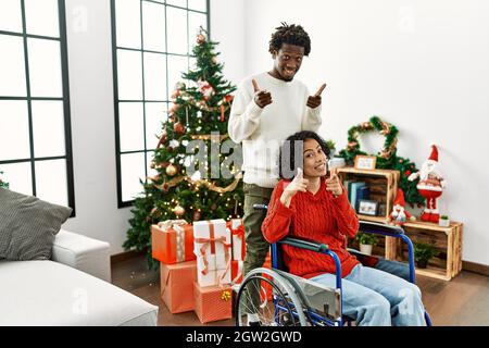 Young interracial couple with woman sitting on wheelchair by christmas tree pointing fingers to camera with happy and funny face. good energy and vibe Stock Photo