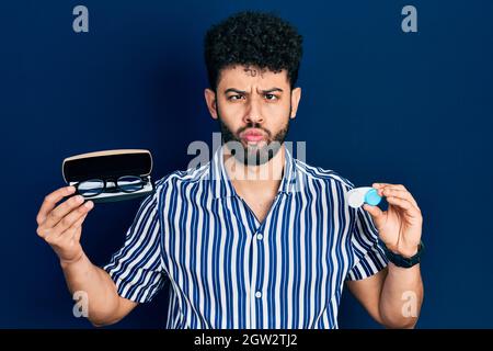 Young arab man with beard holding glasses and contact lenses making fish face with mouth and squinting eyes, crazy and comical. Stock Photo