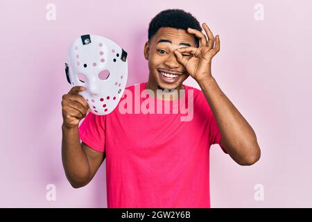 Young african american man holding hockey mask smiling happy doing ok sign with hand on eye looking through fingers Stock Photo