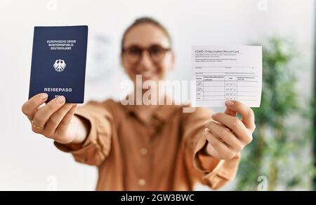 Young blonde woman holding covid vaccination record card and deutschland passport at waiting room Stock Photo