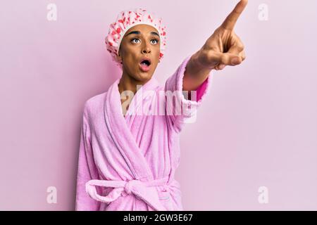 Hispanic man wearing make up wearing shower towel cap and bathrobe pointing with finger surprised ahead, open mouth amazed expression, something on th Stock Photo