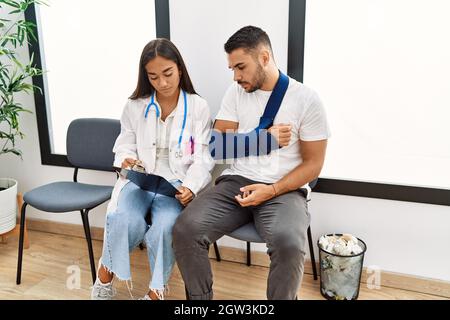 Youg latin injuried man wearing sling talking with the doctor at clinic waiting room. Stock Photo