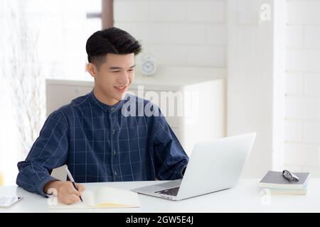 Young asian business man writing on notebook for planning working and using laptop computer on desk at home, notes about finance, male study and learn Stock Photo