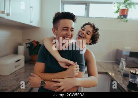 Romantic queer couple embracing each other and laughing together in their kitchen. Happy young queer couple having fun together in the morning. Young Stock Photo