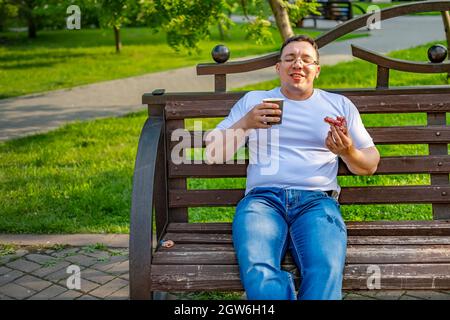 a fat, funny guy with inflated biceps openly enjoys a bun, a donut with coffee, tea, cocoa, sitting on a bench in the park Stock Photo