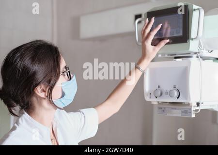 Hospital Radiology Room: Beautiful Multiethnic Woman adjusting X-Ray Machine in Modern Clinic Office. technician in radiology department turning on sc Stock Photo