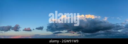 Fluffy cumulus clouds move slowly low over the horizon in the clear blue sky. The top of the cloud lit by the setting sun, and the lower part is dark. Stock Photo