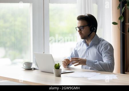 Confident man in wireless headset talking, using laptop, chatting online Stock Photo