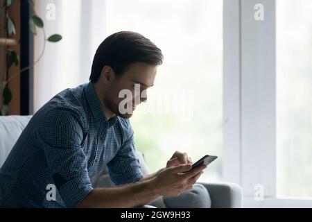 Side view smiling man using smartphone, sitting on cozy couch Stock Photo