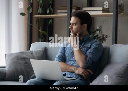 Thoughtful man sitting on couch with laptop, looking in distance Stock Photo