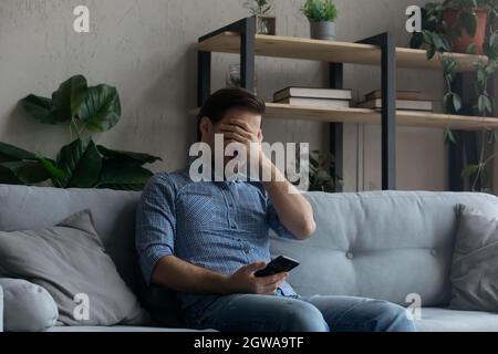 Frustrated young man covering face with hand, holding smartphone Stock Photo