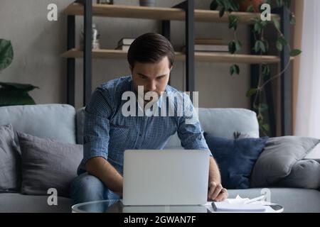 Serious man using laptop, working online, sitting on couch Stock Photo