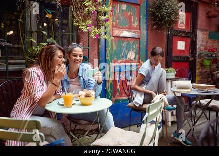 group of people in outdoor cafe. One female friend showing to another something on her cell phone in outdoor cafe. Stock Photo