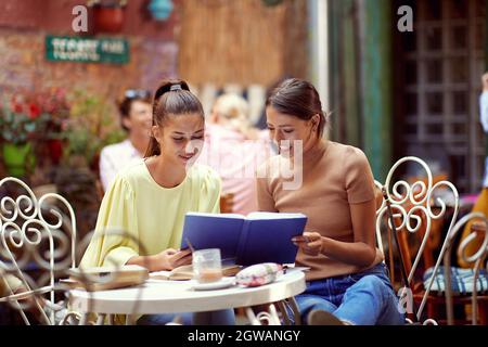 young adult caucasian females reading notebook together, smiling, sitting  in the outdoor cafe Stock Photo