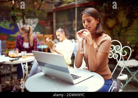 beautiful young caucasian woman sitting in outdoor cafe looking at her laptop on table, thinking. other people in the background Stock Photo