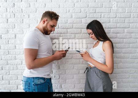 Two friends or lovers standing front of awall and looking to their mobile phones. Technology addiction concept. High quality photo Stock Photo