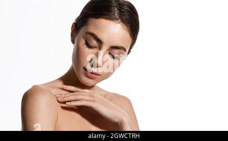 Young woman applying soft skin care cream on her body after shower, gently touching shoulder with fingertips, standing against white background Stock Photo