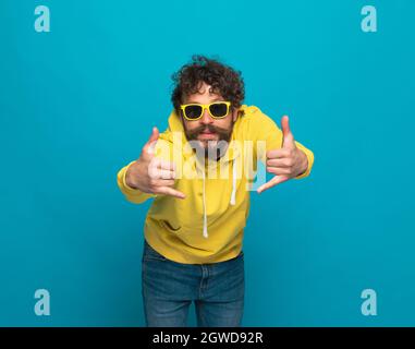 cool bearded man in yellow hoodie making telephone and ringing gestures on blue background in studio Stock Photo