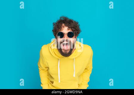 portrait of funny young man in yellow hoodie opening mouth and laughing while posing and standing on blue background in studio Stock Photo