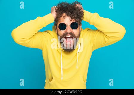 angry young man with glasses and yellow hoodie ripping out hair and screaming while standing on blue background in studio Stock Photo